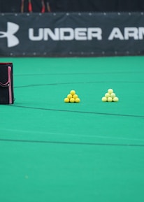 A sports field features a black and pink sports bag, two pyramids of yellow balls, and a portion of a person in dark athletic gear. The background displays a banner with the Under Armour logo.