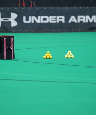 A sports field features a black and pink sports bag, two pyramids of yellow balls, and a portion of a person in dark athletic gear. The background displays a banner with the Under Armour logo.