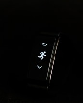 A digital fitness tracker with a black band is displayed against a dark background. The screen features minimalistic white icons for navigation and an exercise mode, including an arrow, a running figure, and a downward-pointing arrow.