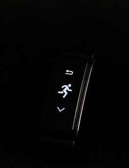 A digital fitness tracker with a black band is displayed against a dark background. The screen features minimalistic white icons for navigation and an exercise mode, including an arrow, a running figure, and a downward-pointing arrow.