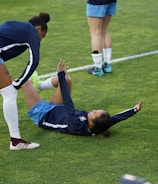 A group of athletes in sportswear are on a green soccer field. Two individuals are interacting closely; one helps the other who is lying on the grass. Nearby, other athletes are stretching.