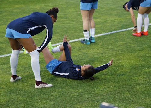 A group of athletes in sportswear are on a green soccer field. Two individuals are interacting closely; one helps the other who is lying on the grass. Nearby, other athletes are stretching.