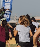 A group of people appears to be participating in an outdoor fitness class or dance session. They are dressed in athletic clothing, and a blue banner can be seen in the background, along with a stage. The setting suggests a lively and energetic atmosphere.