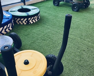 A gym setting with equipment used for weight training resting on artificial grass. There are weighted sleds with handles, featuring colorful plates stacked on them, including yellow and green ones. Additional training apparatuses like large round cushions or bumpers with blue and green covers are visible, designed for strength workouts. The room is bordered by large windows allowing natural light to enter.