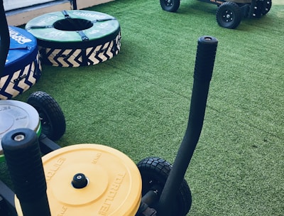 A gym setting with equipment used for weight training resting on artificial grass. There are weighted sleds with handles, featuring colorful plates stacked on them, including yellow and green ones. Additional training apparatuses like large round cushions or bumpers with blue and green covers are visible, designed for strength workouts. The room is bordered by large windows allowing natural light to enter.