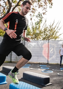 A man is actively participating in an outdoor fitness session, jumping over blue platforms with agility. Two other people in the background are also engaging in exercise, with rings scattered on the ground. The area is fenced and surrounded by trees, suggesting a park or recreational setting.