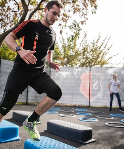 A man is actively participating in an outdoor fitness session, jumping over blue platforms with agility. Two other people in the background are also engaging in exercise, with rings scattered on the ground. The area is fenced and surrounded by trees, suggesting a park or recreational setting.