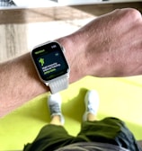 A close-up view of a person's wrist wearing a smartwatch displaying a workout mode for High Intensity Interval Training. The background includes a bright green exercise mat on a wooden floor. The person is wearing white athletic shoes and dark shorts.
