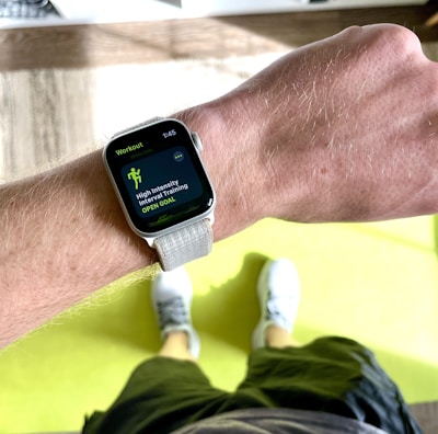 A close-up view of a person's wrist wearing a smartwatch displaying a workout mode for High Intensity Interval Training. The background includes a bright green exercise mat on a wooden floor. The person is wearing white athletic shoes and dark shorts.