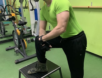 A man wearing a bright green athletic shirt, black pants, and a cap is standing in a gym. He has one foot on a small platform and is smiling at the camera. In the background, there are exercise bikes and artwork on the green walls.