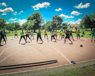 A group of people are exercising outdoors on a concrete court, surrounded by green spaces and trees. They are standing in a formation, appearing to engage in an organized fitness class. Each participant wears athletic clothing, and several carry exercise equipment such as dumbbells and ropes. The sky is bright blue with scattered fluffy clouds, creating a vibrant and energetic atmosphere.