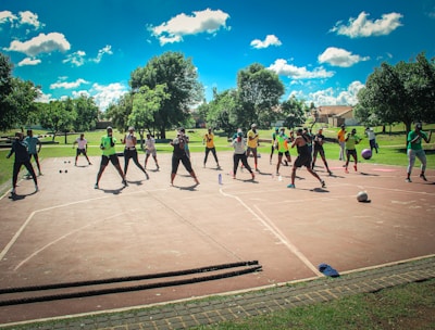 A group of people are exercising outdoors on a concrete court, surrounded by green spaces and trees. They are standing in a formation, appearing to engage in an organized fitness class. Each participant wears athletic clothing, and several carry exercise equipment such as dumbbells and ropes. The sky is bright blue with scattered fluffy clouds, creating a vibrant and energetic atmosphere.