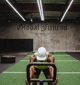 A person in athletic wear is pushing a sled on green turf inside a dimly lit gym with 'Global Fitness' written on the wall. The setting includes exercise equipment and industrial elements, such as visible metal beams and a rolled-up garage door.