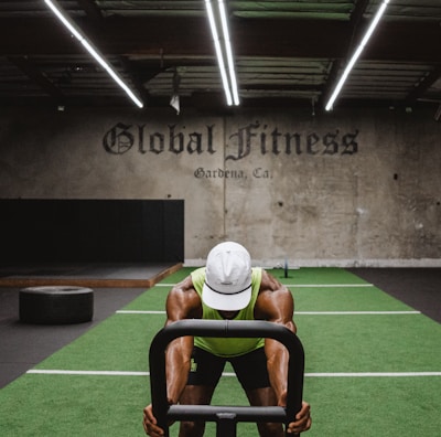 A person in athletic wear is pushing a sled on green turf inside a dimly lit gym with 'Global Fitness' written on the wall. The setting includes exercise equipment and industrial elements, such as visible metal beams and a rolled-up garage door.