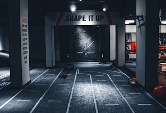 A dimly lit indoor gym with a central pathway marked with lane lines. The ceiling features multiple lights, and there are exercise equipment and motivational signage, including one that reads 'SHAPE IT UP'. The ambiance suggests a focus area for fitness activities.