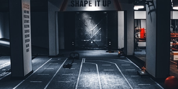 A dimly lit indoor gym with a central pathway marked with lane lines. The ceiling features multiple lights, and there are exercise equipment and motivational signage, including one that reads 'SHAPE IT UP'. The ambiance suggests a focus area for fitness activities.