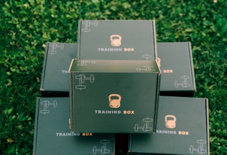 Several black boxes labeled 'Training Box' are stacked on a grassy surface. The top box features illustrations of a kettlebell and a dumbbell.
