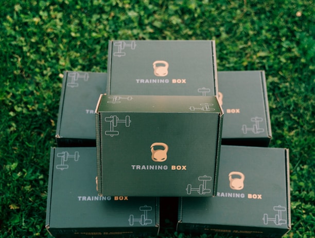 Several black boxes labeled 'Training Box' are stacked on a grassy surface. The top box features illustrations of a kettlebell and a dumbbell.