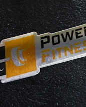 A metal plaque with the text 'POWER FITNESS' is set against a textured black background. The design includes a stylized dumbbell icon adjacent to the text, with a modern and sleek appearance.