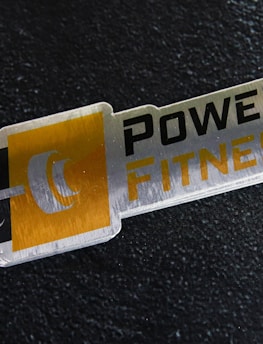 A metal plaque with the text 'POWER FITNESS' is set against a textured black background. The design includes a stylized dumbbell icon adjacent to the text, with a modern and sleek appearance.
