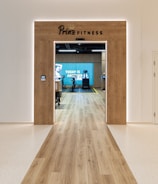 A modern fitness center entrance with wooden framing and the sign 'PRIME FITNESS' above the doorway. The interior features a spacious gym area with workout equipment visible and a motivational banner on the back wall. The flooring transitions from glossy tiles to a wood finish leading into the facility.