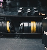 A gym setting featuring various fitness equipment, including kettlebells, stacked weight plates on a rack, dumbbells, and a medicine ball. The gym has a rugged, industrial vibe with black walls and a Bulldog Gear plyo box.