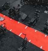 A gym setting featuring a variety of exercise equipment such as weight machines, stationary bikes, and a sled on a bright red track. The gym has a modern aesthetic with black and red color schemes.