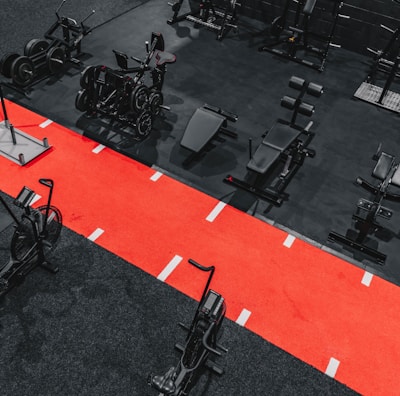 A gym setting featuring a variety of exercise equipment such as weight machines, stationary bikes, and a sled on a bright red track. The gym has a modern aesthetic with black and red color schemes.