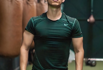 A person is standing confidently in a gym-like setting, wearing a dark green athletic shirt and light brown shorts. Several punching bags are visible hanging in the background, suggesting a boxing or fitness environment.