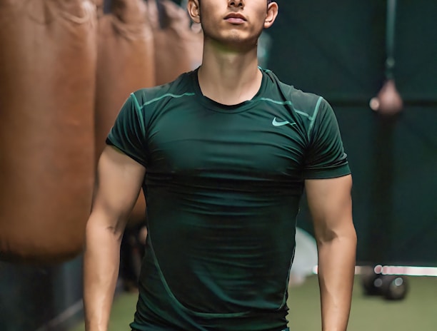 A person is standing confidently in a gym-like setting, wearing a dark green athletic shirt and light brown shorts. Several punching bags are visible hanging in the background, suggesting a boxing or fitness environment.