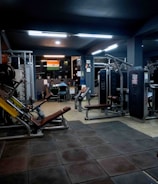 A dimly lit gym with various exercise machines, including leg presses and cable equipment. The floor is padded with dark tiles. In the background, two people are sitting at a desk with a computer. An Indian flag is visible on the wall, adding a touch of decoration.