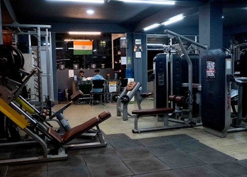 A dimly lit gym with various exercise machines, including leg presses and cable equipment. The floor is padded with dark tiles. In the background, two people are sitting at a desk with a computer. An Indian flag is visible on the wall, adding a touch of decoration.