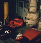 A dimly lit gym or training room featuring a pair of red boxing gloves resting on a surface. The background includes a wrapped pillar and a boxing bag, suggesting an atmosphere of sports training.