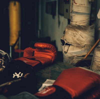 A dimly lit gym or training room featuring a pair of red boxing gloves resting on a surface. The background includes a wrapped pillar and a boxing bag, suggesting an atmosphere of sports training.