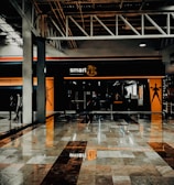 A fitness center entrance with the branding 'smart fit' illuminated above the doors. The interior has a modern design, featuring dark glass and bright orange accents. A reflective marble floor extends in front, capturing reflections from the surroundings. There are two individuals standing and walking towards the left.