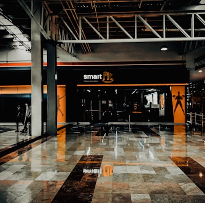 A fitness center entrance with the branding 'smart fit' illuminated above the doors. The interior has a modern design, featuring dark glass and bright orange accents. A reflective marble floor extends in front, capturing reflections from the surroundings. There are two individuals standing and walking towards the left.